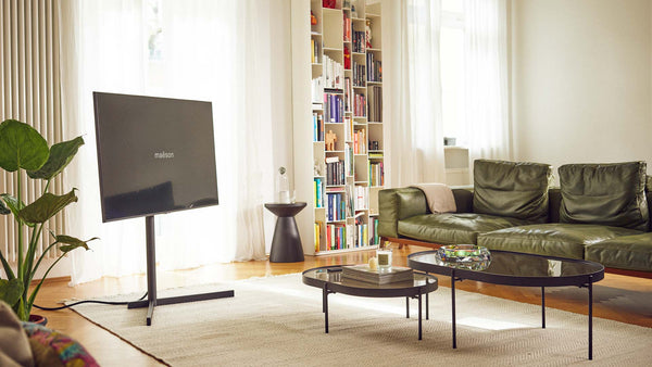 Drehbarer TV Standfuß AURA mit Fernseher im Wohnzimmer - frei auf dem Teppich vor Bücherregal und Sofa platziert